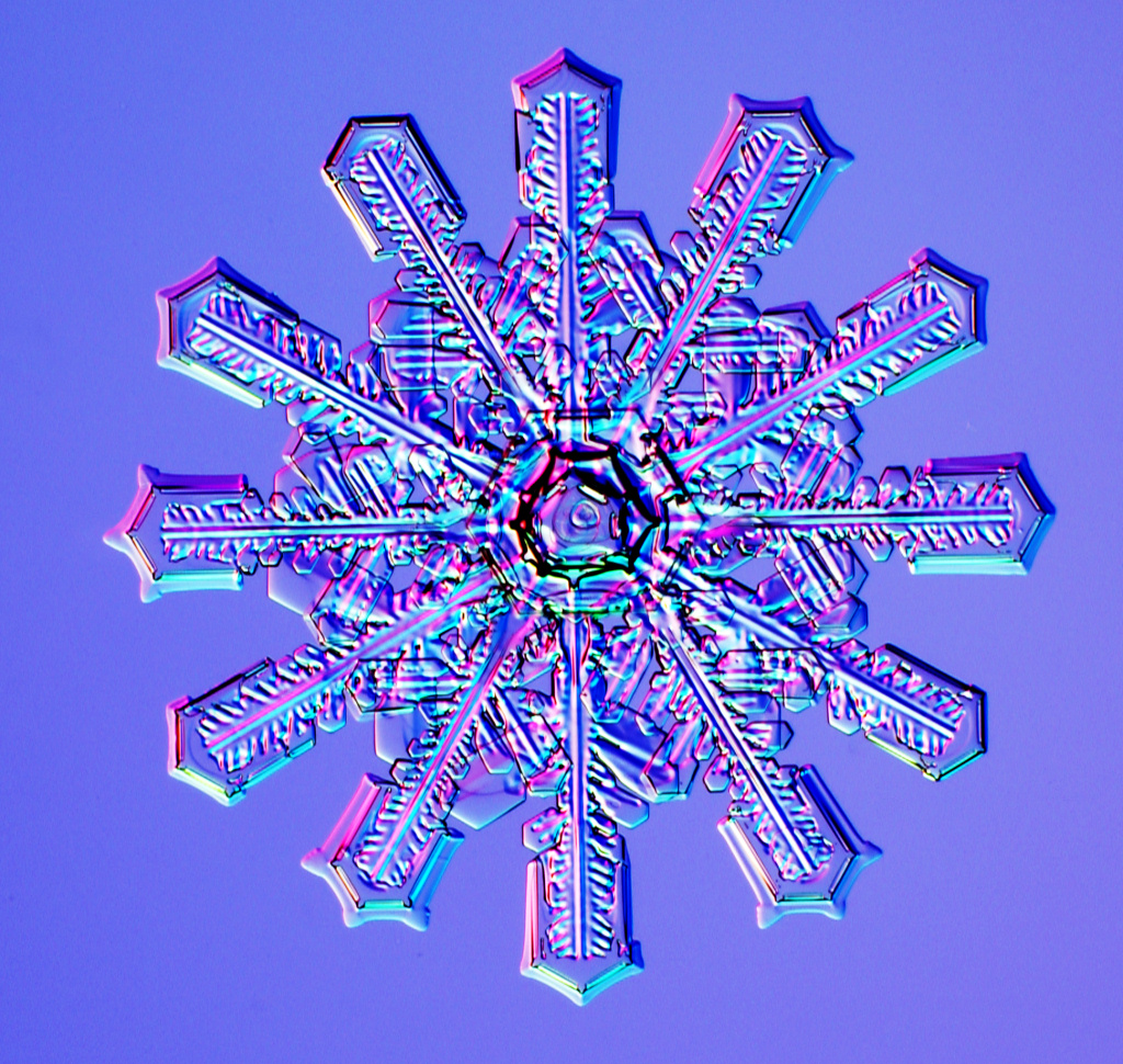 This is an image of a twelve-branched snowflake. The branches radiate around the hexagonal centre like the hands of a clock. The snowflake is placed on a translucent blue-mauve background and the crystalline branches reflect hues of pink and light blue.