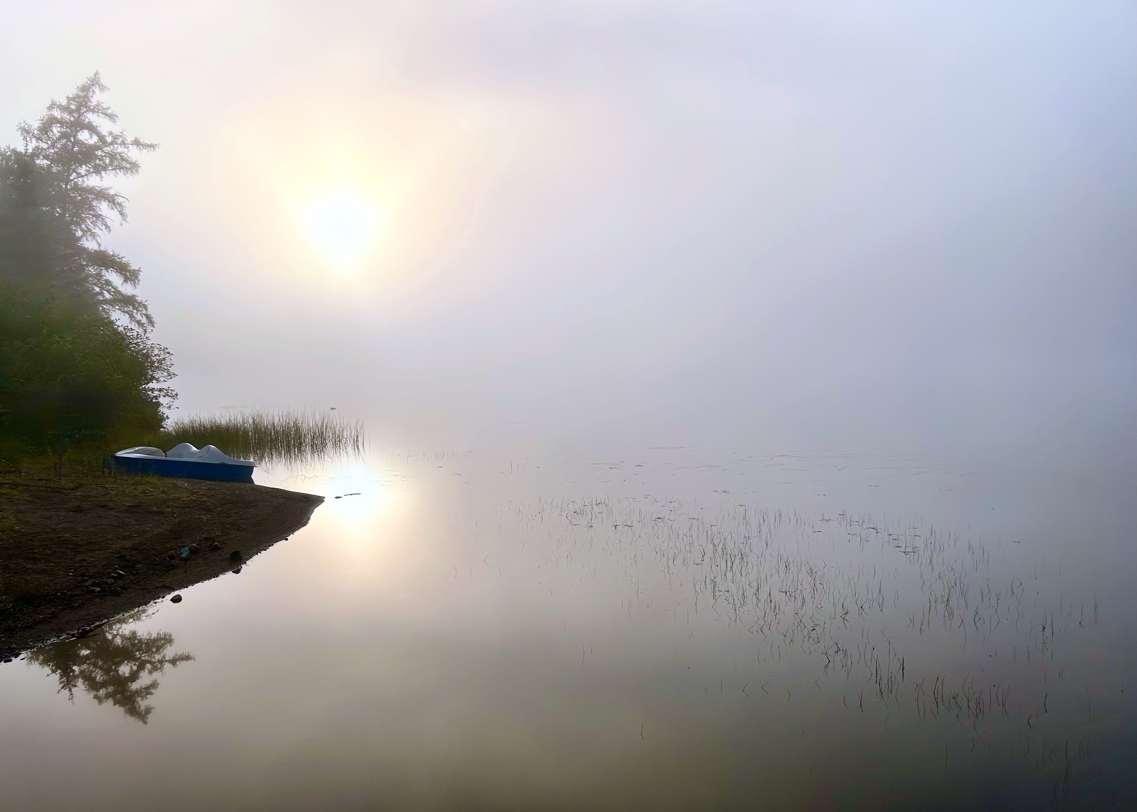 This is a photomontage depicting a cloud of unknowing. On the left of the image is the beach of a lake, with a boat on a point of sand. The shoreline extends in a sharp line, but the water merges into mist that covers most of the right side of the image. A low sun, just above the boat, is reflected in the water by the shore, suggesting that the clouds will lift and clarity will be unveiled.